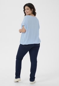 Haut bleu clair à manches courtes avec une coupe décontractée, associé à un jean bleu foncé. Le modèle porte des baskets beiges texturées. Vue de dos, fond neutre.