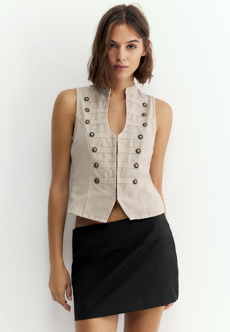 Jeune femme vêtue d'un gilet beige sans manches avec des boutons et d'une mini-jupe asymétrique noire, debout devant un fond blanc uni.