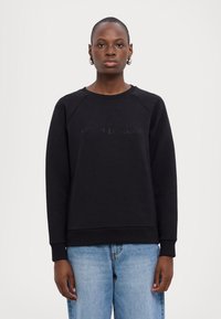 Felpa nera con maniche lunghe a raglan e scollatura a girocollo, con logo testurizzato sul petto. Abbinata a un paio di jeans azzurri chiari.
