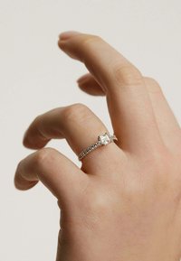 Anillo de plata con una piedra central de corte esmeralda y una banda con pequeñas piedras preciosas claras, llevado en una mano de tono de piel claro.