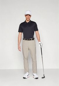 Ținută de golf pentru bărbați, formată dintr-un polo negru mulat, pantaloni bej, adidași albi și o șapcă albă. Ținând un croșet de golf, în picioare, drept.