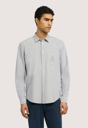 Mann mit lockigem dunklem Haar, der ein hellgraues Hemd mit Brusttasche und dunkelblaue Jeans trägt und leicht zur Seite blickt.