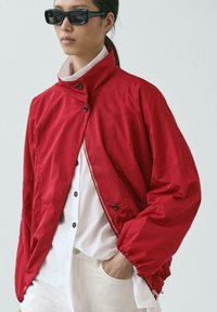 Personne portant des lunettes de soleil foncées, une veste ample rouge vif par-dessus une chemise blanche à boutons, et un pantalon blanc, avec une main dans la poche.