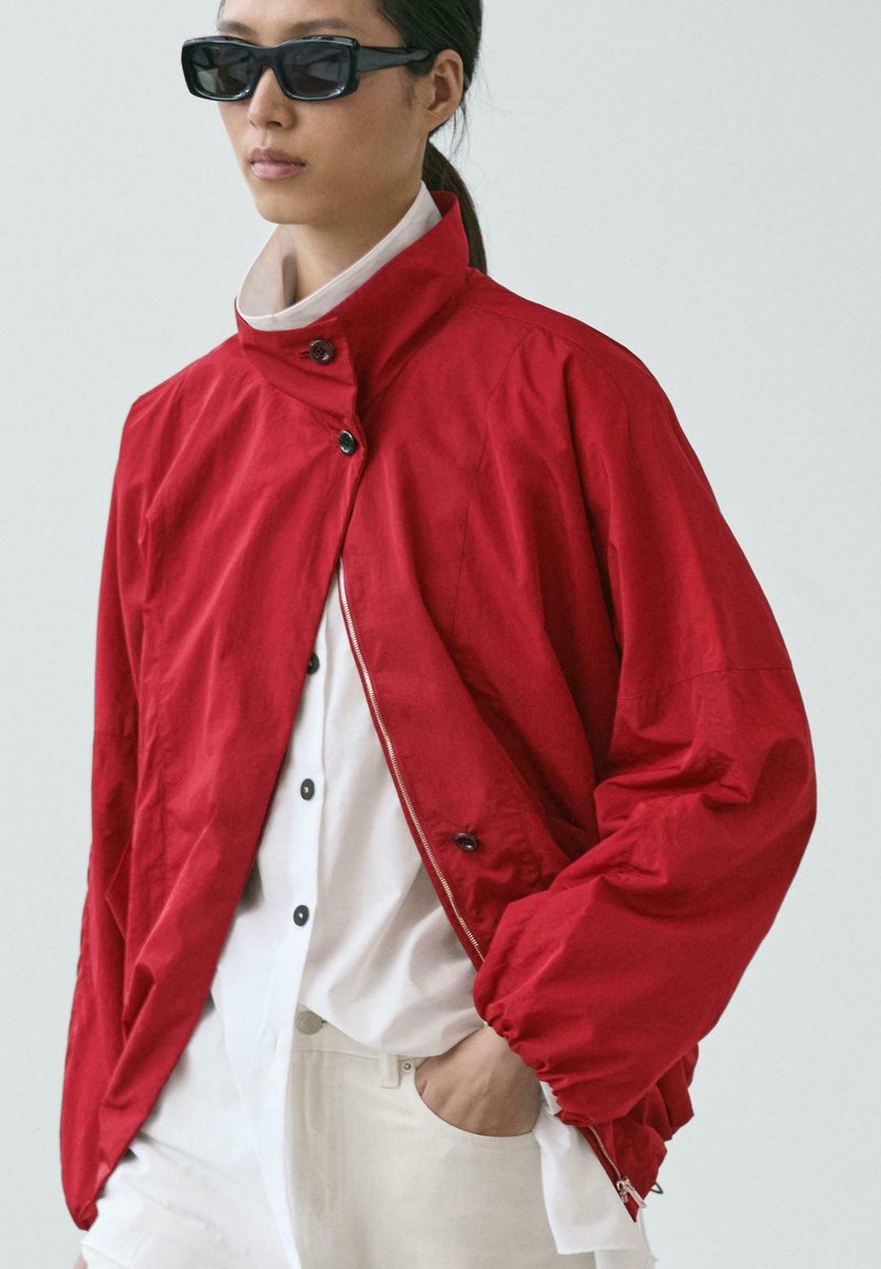 Personne portant des lunettes de soleil foncées, une veste ample rouge vif par-dessus une chemise blanche à boutons, et un pantalon blanc, avec une main dans la poche.