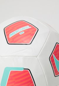 Valkoinen jalkapallo, jossa on kuusikulmaisia paneeleja, punaruskeareunuksinen muotoilu ja "Mercurial"-logo turkoosina. Pinta on sileä ja saumat ommeltu.