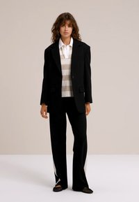 Melnas uzvalka jakas virs balta svītrainā krekla un melnām biksēm ar sānu šķēlumiem, ar kontrastējošiem gaišiem akcentiem un plakanām kurpēm.