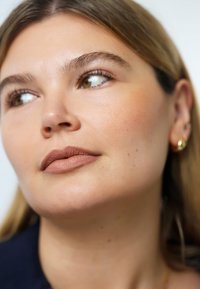 Close-up van een gezicht met een lichte huidtint, neutrale lipkleur en subtiele highlighter op de jukbeenderen. Kenmerken zijn onder andere bruine ogen en minimale sieraden.