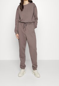 Quiltad brun tröja och matchande byxor i mjukt tyg, med snörning i midjan och elastiska mudd, kombinerat med ljusa stövlar.