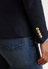 Blazer azul-marinho texturizado com botões dourados e punho às riscas azuis visível. Combinado com jeans denim que mostram um detalhe do bolso traseiro.