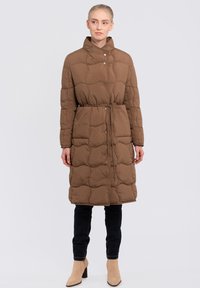 Brown quilted puffer coat with a high collar, snap buttons, adjustable waist tie, and long sleeves. Worn over dark pants and beige ankle boots.