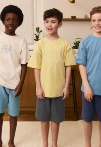 Tre børn står sammen iført løstsiddende t-shirts i creme, gul og lyseblå, kombineret med shorts i grå og blå. Tekstdetaljer på skjorterne.