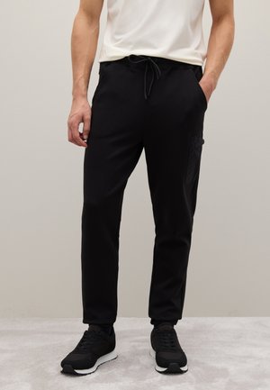 Pantaloni da jogging neri con vita regolabile, tasche laterali e dettaglio cargo testurizzato sulla gamba sinistra. Indossati con sneakers nere.