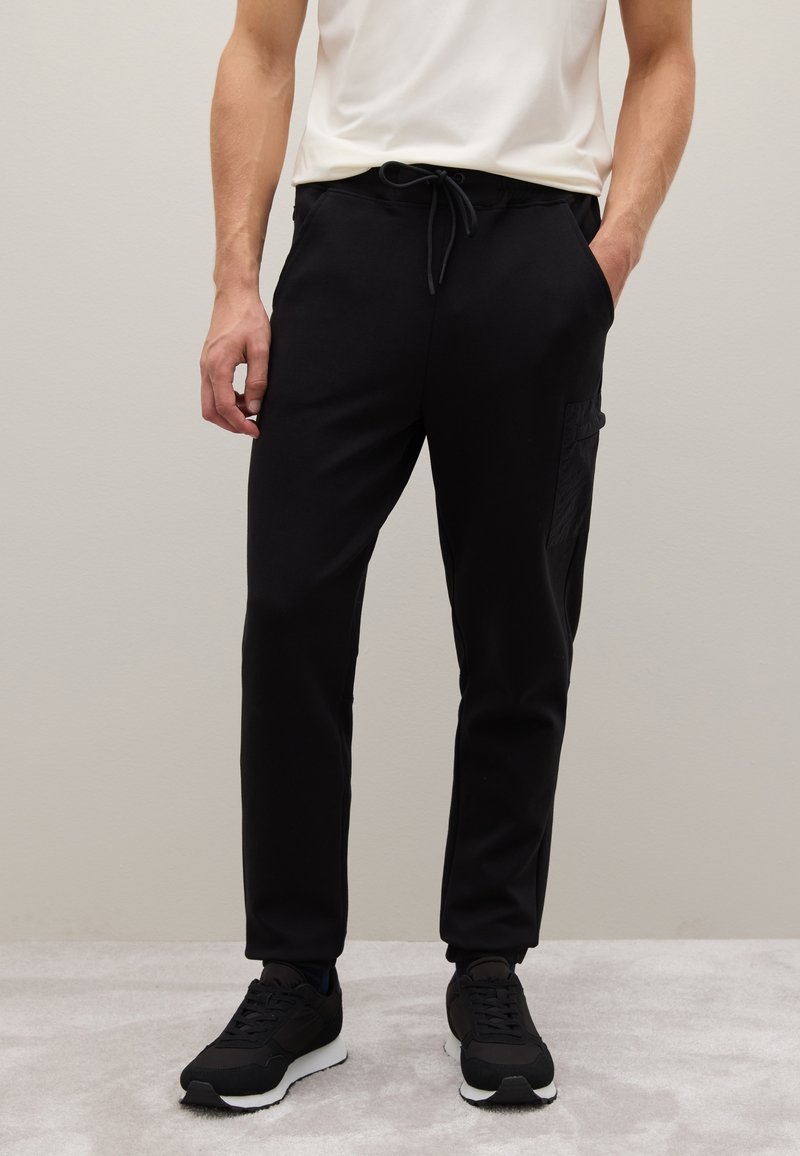 Schwarze Jogginghosen mit einem Kordelzugbund, seitlichen Taschen und einem strukturierten Cargo-Taschen-Detail am linken Bein. Getragen mit schwarzen Sneakers.