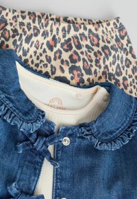 Giubbotto di jeans con colletto volante, di colore blu, indossato sopra una maglietta a coste beige. Leggings con stampa animale con macchie pesca e nere sotto.