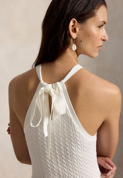 Kvinne med langt mørkt hår iført en hvit, teksturert ermeløs topp bundet med sløyfe bak, sett fra siden, med ørepynt formet som en skjell.