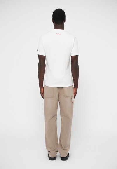 Λευκό βαμβακερό μπλουζάκι με κοντά μανίκια, με κόκκινο κείμενο στην πλάτη, συνδυασμένο με μπεζ φαρδιά παντελόνια και μαύρα παπούτσια.