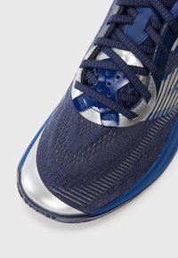 Close-up of a navy blue running shoe with silver toe cap, detailed knit pattern, and navy blue laces threaded through plastic eyelets.