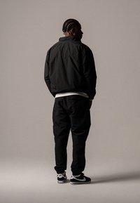 Jachetă neagră ușoară cu manșete elastice, purtată peste o cămașă albă, asociată cu pantaloni negri tapered și adidași negri Nike, văzută din spate.