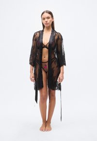 Μαύρο κιμονό από δαντέλα με κεντήματα λουλουδιών, διαφανές ύφασμα, ανοιχτό μπροστά, μανίκια τριών τετάρτων, σατέν κορδόνια και τελείωμα με κυματιστό σχέδιο. Λεπτή, με λουριά εσωρούχα από κάτω.