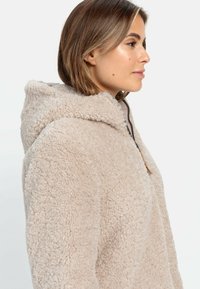 Beige Fleece-Hoodie mit strukturierter Oberfläche, ausgestattet mit einer Kapuze und einem Frontreißverschluss. Keine sichtbaren Muster oder Hardwaredetails.