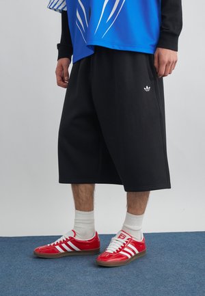 Muškarac nosi crne Adidas kratke hlače do koljena, crvene Adidas tenisice s bijelim prugama, bijele čarape i plavu košulju s uzorkom.