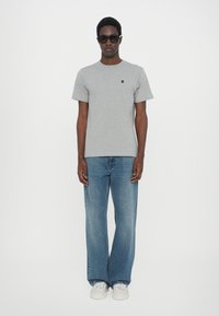 Tricou gri cu mânecă scurtă și un logo mic, asortat cu blugi largi de culoare albastru deschis și adidași albi, purtat de un model masculin în picioare.