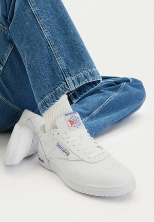 Białe trampki Reebok z niebiesko-czerwonym logo, noszone z białymi skarpetkami i niebieskimi dżinsami na gładkim białym tle.