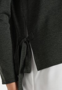 Pull en maille vert foncé avec une texture douce, présentant un détail de nœud sur le côté et une coupe décontractée. Des manches longues et un ourlet arrondi complètent le design.