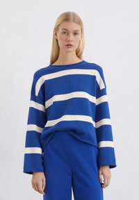 Blauer gestreifter Pullover mit breiten weißen Streifen, lockere Passform, runder Halsausschnitt und langen Ärmeln. Kombiniert mit passenden blauen Hosen.