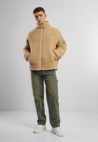 Jachetă din fleece bej cu guler înalt, închidere cu fermoar și buzunare laterale, asociată cu blugi verde deschis și adidași albi.