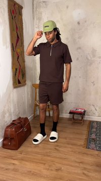 Hombre con camisa marrón y pantalones cortos, con calcetines negros y sandalias blancas, ajustándose una gorra verde claro en una habitación con suelo de madera y pared texturizada.