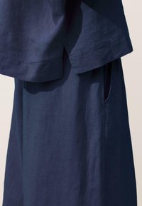 Tengerészkék lenvászon ruhadarab átfedő réteges szegéllyel és oldalsó hasítékos zsebbel, amely megmutatja a textúra és varrás részleteit.