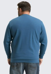 Niebieska bluza z długimi rękawami, ściągaczami przy mankietach i dole oraz gładką teksturą. Noszona z ciemnymi dżinsami, które mają kontrastowe szwy.