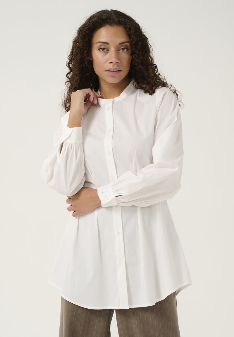 Camicia bianca con colletto rotondo, frontale plissettato e maniche lunghe a sbuffo. Realizzata in tessuto morbido con vestibilità rilassata e orlo curvo.