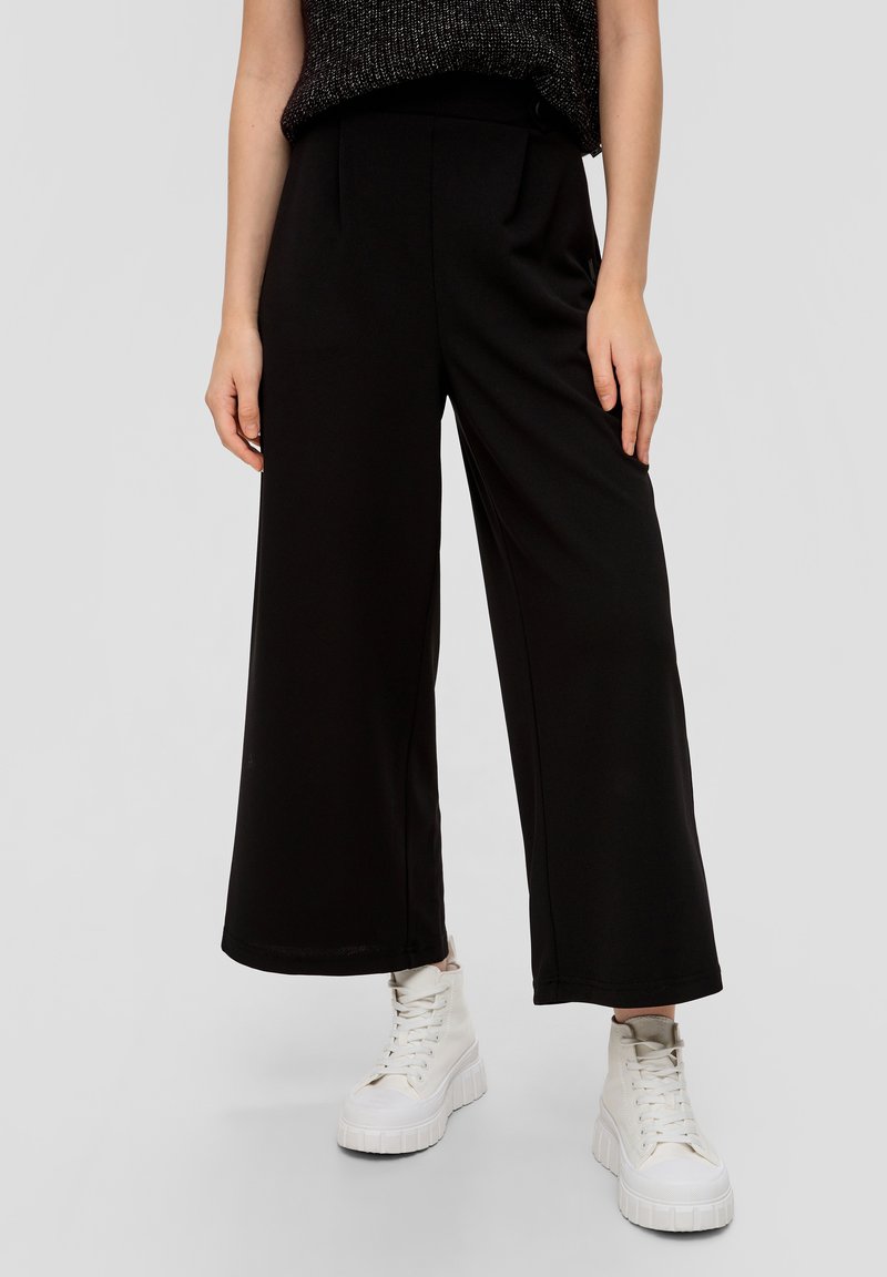 Pantaloni neri a gamba larga con un design sartoriale, caratterizzati da pieghe sul davanti, realizzati in un tessuto liscio, abbinati a sneakers bianche chunky.