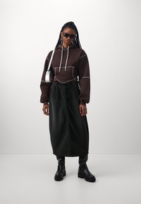 Sudadera corta marrón con costuras blancas, combinada con una falda maxi de mezclilla negra y botas negras de tobillo. Accesorio: pequeño bolso de hombro metálico.