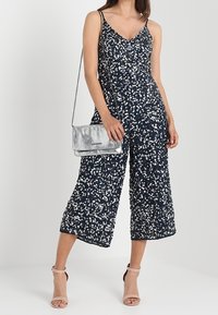 Tuta floreale blu navy con scollo a V, spalline sottili e pantaloni a zampa d'elefante, abbinata a una pochette metallica argento e tacchi color nudo.