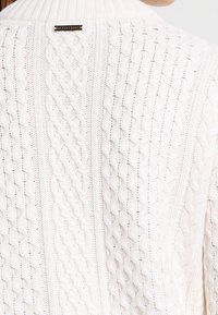 Weißer Strickpullover mit hohem Kragen, strukturiertem Muster und einem kleinen metallischen Logoschild auf der Rückseite nahe dem Kragen.