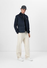 Marineblå quiltet vest med glidelås i front, to frontlommer, sammen med en svart turtleneck, kremfargede vide bukser og hvite sneakers.