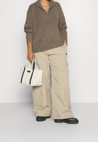 Pull marron en tricot avec une demi-fermeture éclair, pantalon cargo beige ample, et un petit sac en fausse fourrure blanche avec des accents noirs et des poignées. Bottes à plateforme noires.