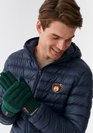 Guanti verdi a maglia con polsini a costine, caratterizzati da un piccolo dettaglio di ricamo a forma di orsetto. Indossati da una persona con un giubbotto imbottito blu navy con un patch a forma di orso.