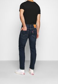 Man wearing dark blue jeans, black t-shirt, and white high-top sneakers standing with one hand in back pocket facing away