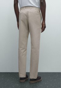Beige trousers with a slim fit, featuring two back pockets and a smooth, textured fabric. Worn with dark shoes on a patterned surface.