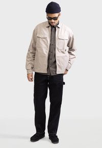 Beige canvas jacket with two chest pockets, worn over a brown checkered shirt and paired with black pants and shoes. Black knit hat and sunglasses.