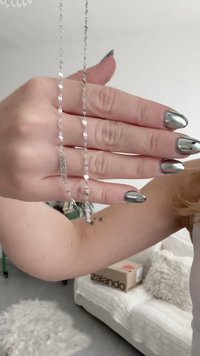 Ruka sa srebrnim metalik noktima drži srebrnu ogrlicu s lancem na kojem je ugravirano "GUESS"; u pozadini se vidi sofa i paket Zalando.