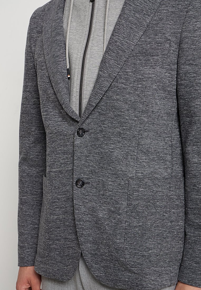 Szary żakiet wykonany z teksturowanej tkaniny, z kołnierzem typu szal, dwoma przednimi kieszeniami i podwójnym zapięciem na guziki. Noszony na szarym hoodie.