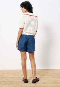 Strickpullover mit rotem Rand, kombiniert mit Jeansshorts. Der Pullover ist weiß mit offenem Muster, und die Shorts sind dunkelblau und gehen bis zur Mitte des Oberschenkels.