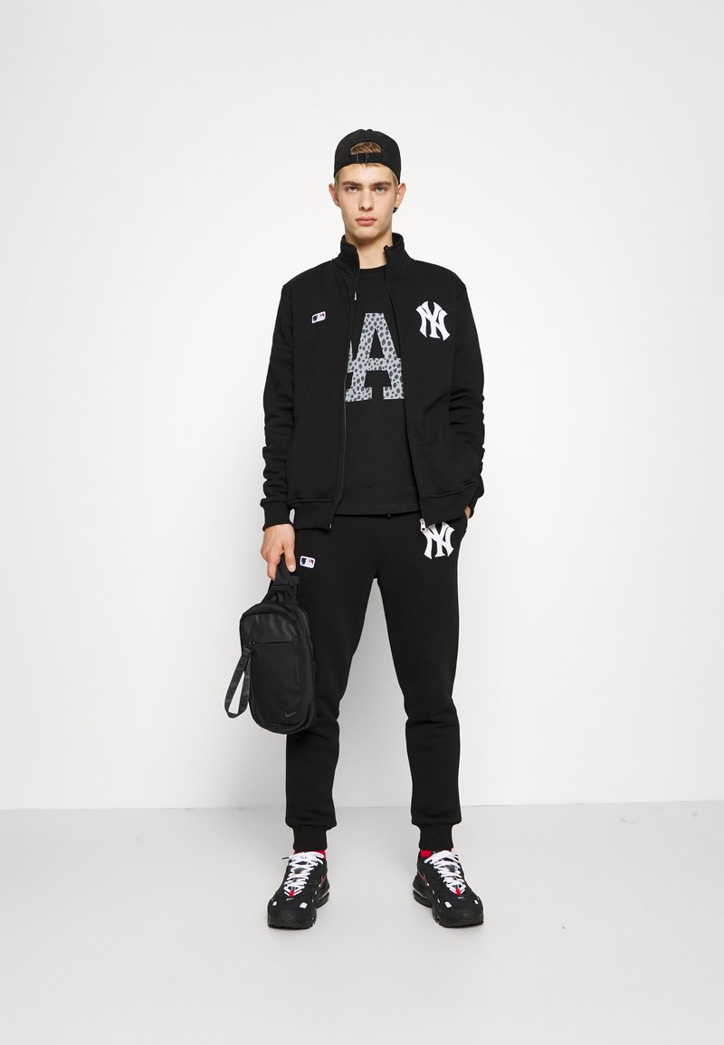 Black athletic outfit including a zip-up jacket, graphic t-shirt with an emblem, and jogger pants. Accessories include a black bag and sneakers.