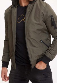 Giacca bomber verde oliva con zip frontale, polsini a costine e tasche laterali. Presenta un cappuccio e tessuto testurizzato. Indossata sopra una maglietta nera.