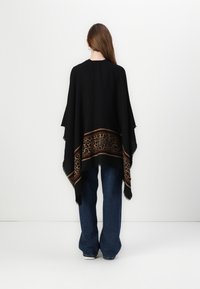 Poncho nero con bordo a stampa leopardata e dettagli marroni, caratterizzato da un orlo frangiato e un design fluido, indossato sopra jeans a zampa blu.